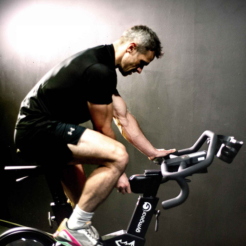 Man exercising on the Gym Gear M Sport Elite Indoor Cycle exercise bike against a dark background