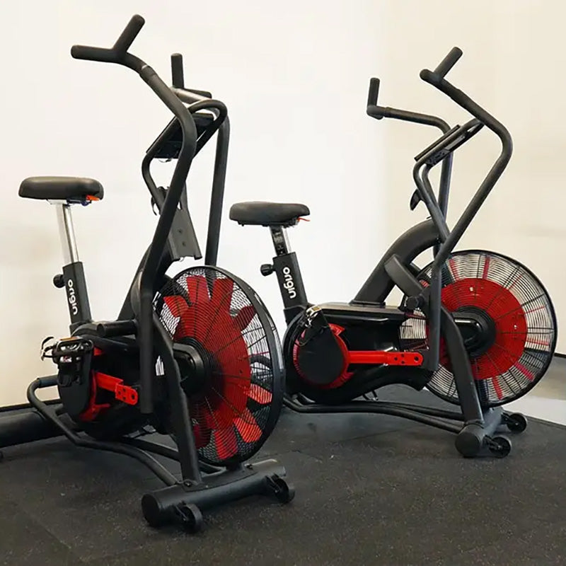 Two Origin Storm Air Bike exercise bike with red fans on a black floor against a white wall.