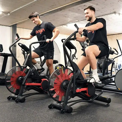Two men exercising on the Origin Storm Air Bike exercise bike in a gym setting.