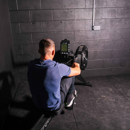 Person using the Gym gear Row Max 2.0 rowing machine in a dark room with grey tiled walls.