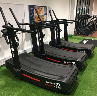 Row of Gym Gear Curve 2.0 PLUS Treadmill in black on a green floor with a white wall in the background.
